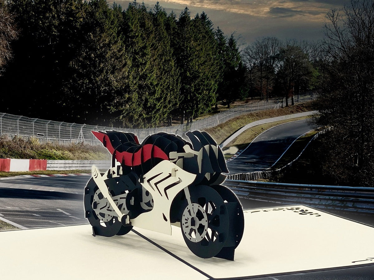 Pop-up-Karte mit Rennmotorrad, aufgestellt vor einer echten Rennstrecke mit Leitplanken und Wald.