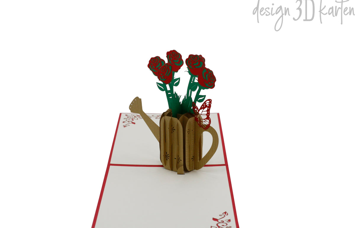Aufgeklappte Pop-up-Karte mit roten Rosen in einer goldenen Gießkanne, frontal fotografiert.