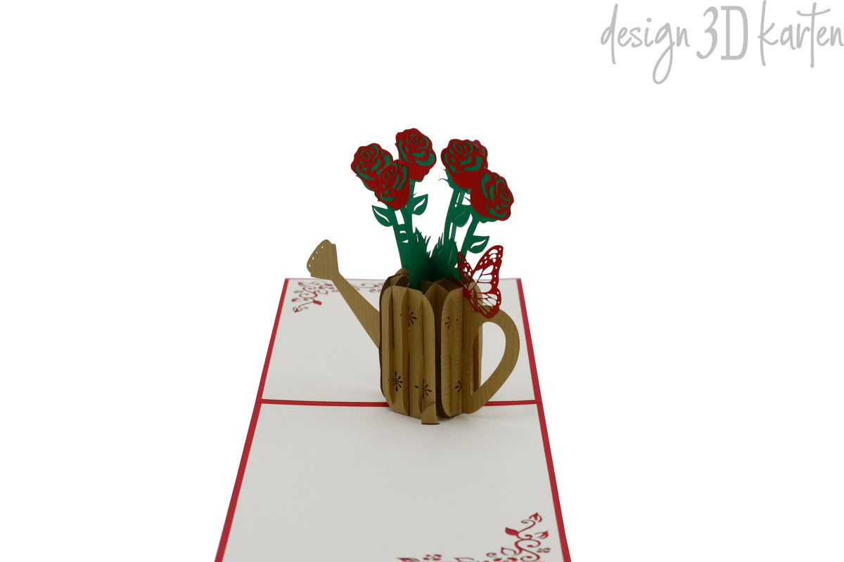 Aufgeklappte Pop-up-Karte mit roten Rosen in einer goldenen Gießkanne, frontal fotografiert.