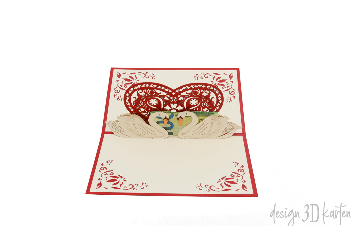 Aufgeklappte Pop-up-Karte mit Schwanenpaar unter einem roten Ornamentherz, schräg von der Seite fotografiert.
