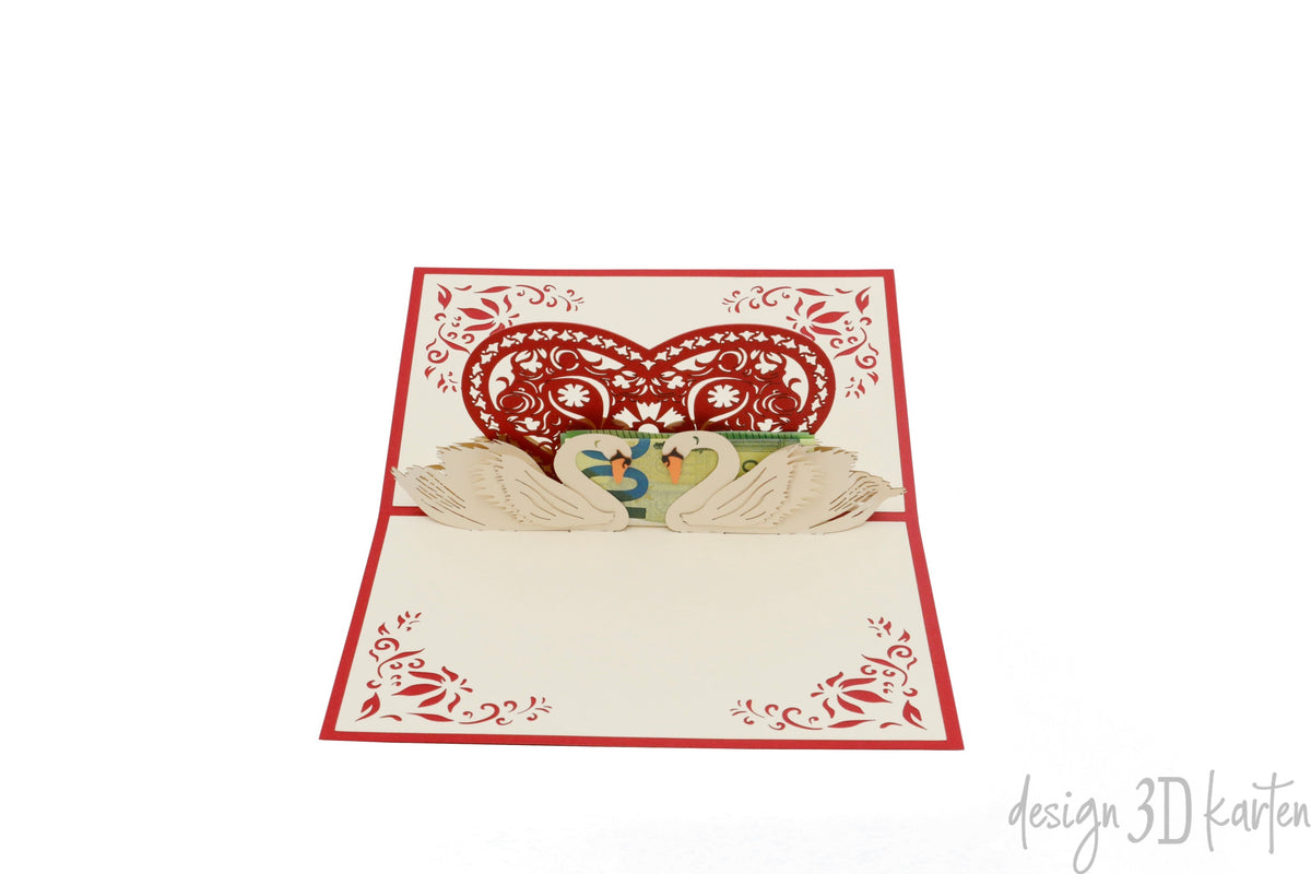 Aufgeklappte Pop-up-Karte mit Schwanenpaar unter einem roten Ornamentherz, schräg von der Seite fotografiert.
