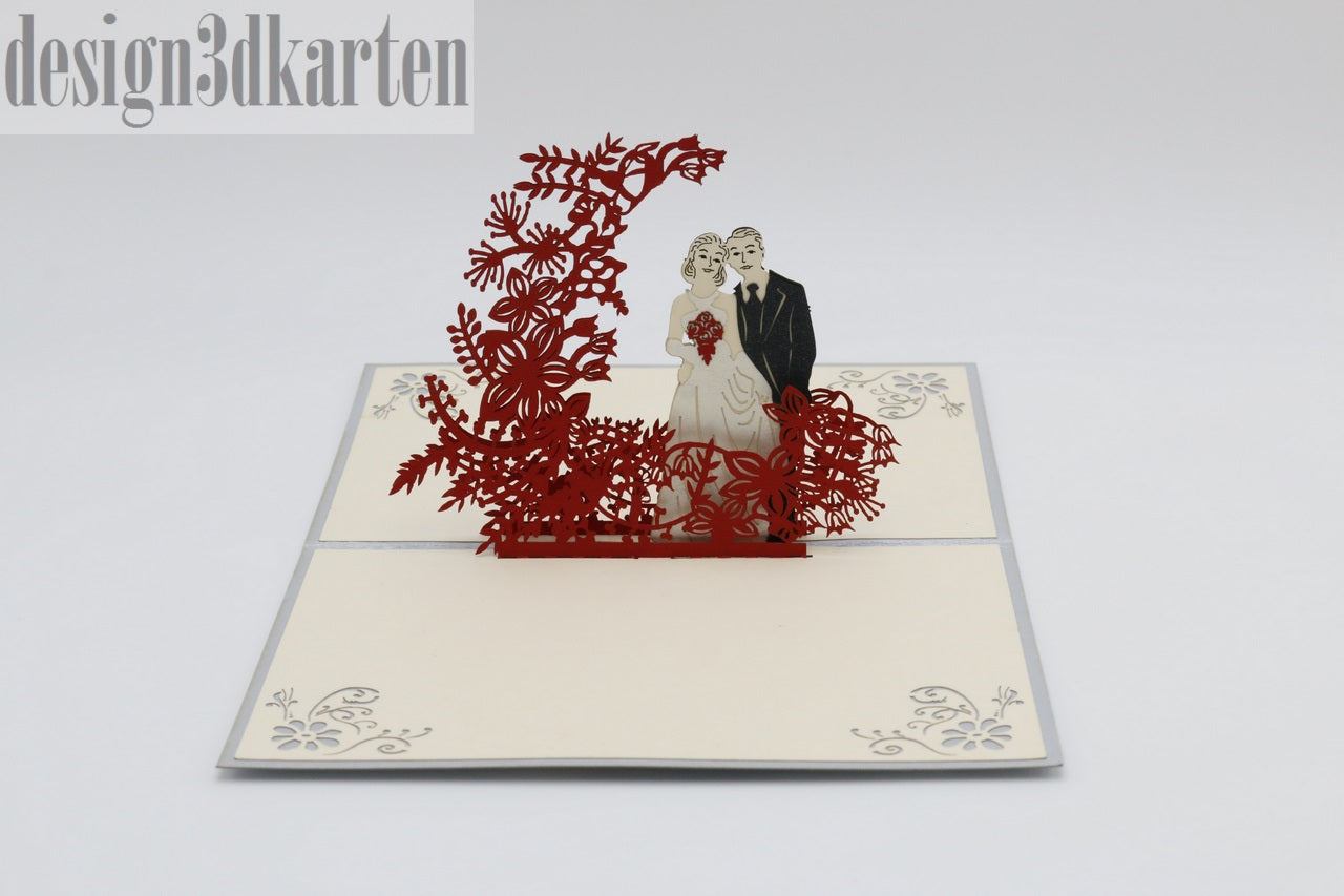 Aufgeklappte Pop-up-Karte mit Brautpaar unter einem roten Blütenbogen, schräg von oben fotografiert.