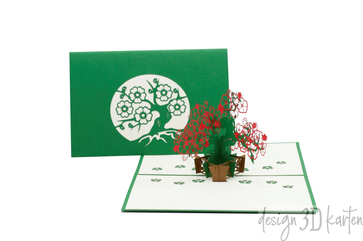 Aufgeklappte Pop-up-Karte mit blühendem Blumenbonsai, dahinter die grüne Kartenvorderseite mit dem Bonsai-Scherenschnitt.