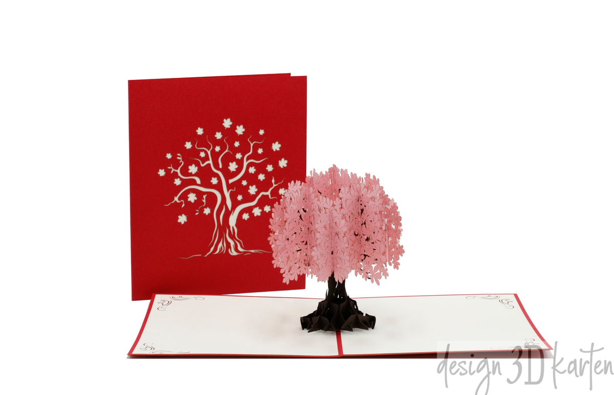 Aufgeklappte Pop-up-Karte mit einem rosa Blütenbaum, daneben die rote Kartenvorderseite mit Baum-Scherenschnitt.