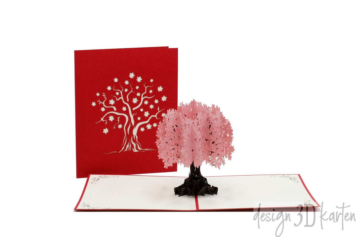 Aufgeklappte Pop-up-Karte mit einem rosa Blütenbaum, daneben die rote Kartenvorderseite mit Baum-Scherenschnitt.