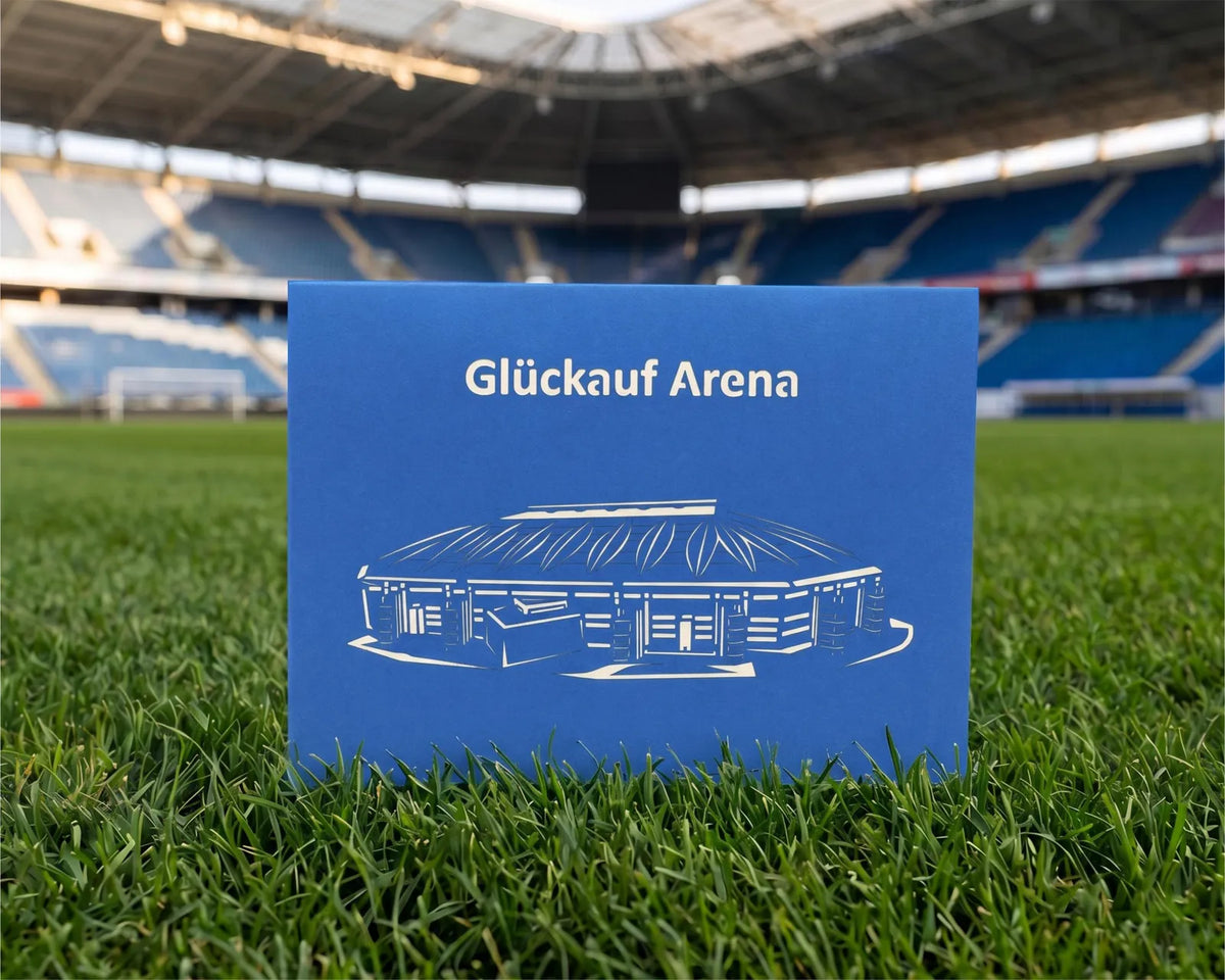 Blaue Klappkarte mit Scherenschnitt eines Fußballstadions und dem Schriftzug Glück auf Arena auf Rasen im Stadion.