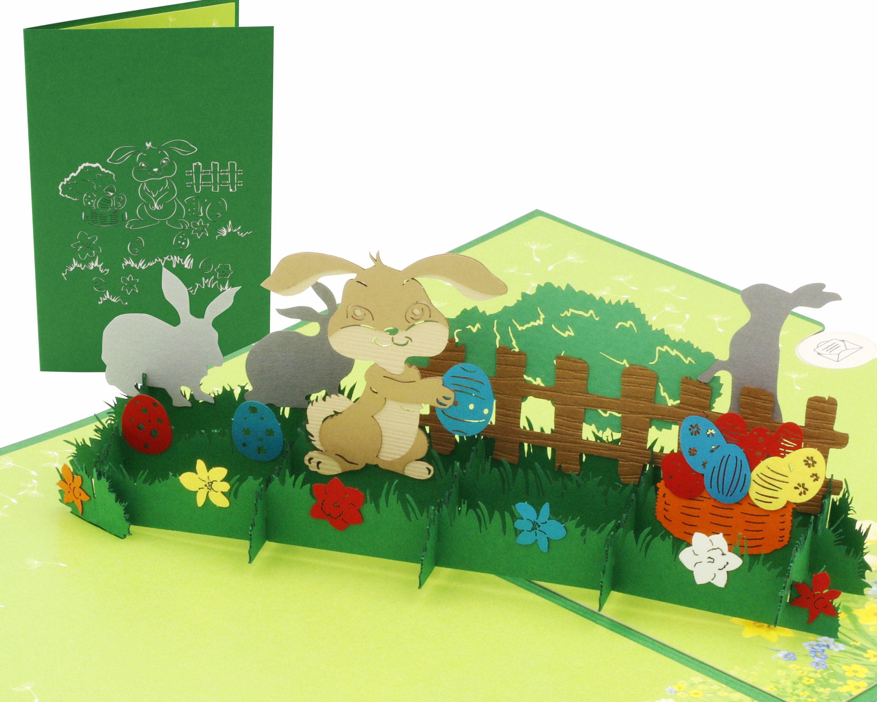 Aufgeklappte Pop-up-Karte mit Osterhasen, Ostereiern und Gartenzaun auf einer Frühlingswiese, daneben die grüne Kartenvorderseite mit Hasen-Scherenschnitt.