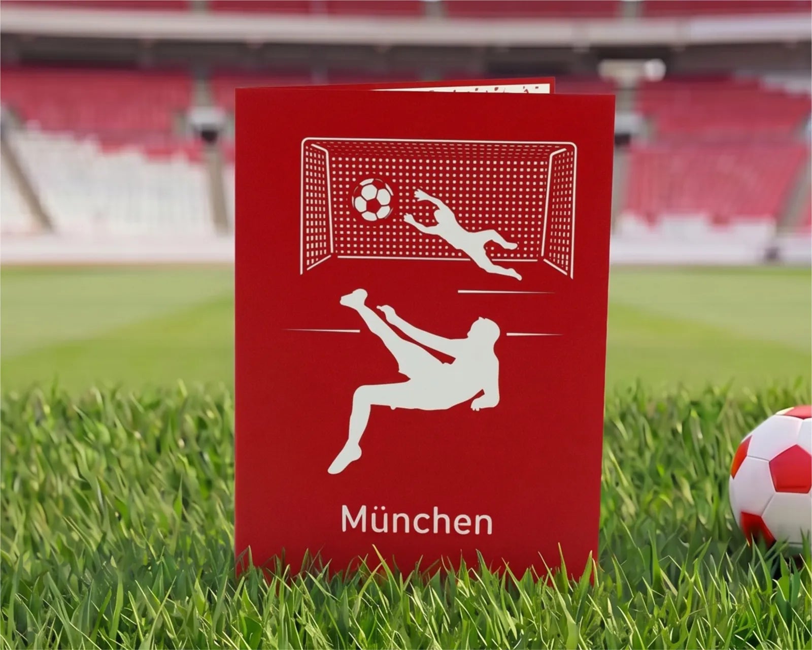 Rote Klappkarte mit Scherenschnitt eines Fußballspielers beim Fallrückzieher vor einem Tor und dem Schriftzug München auf Rasen im Stadion.