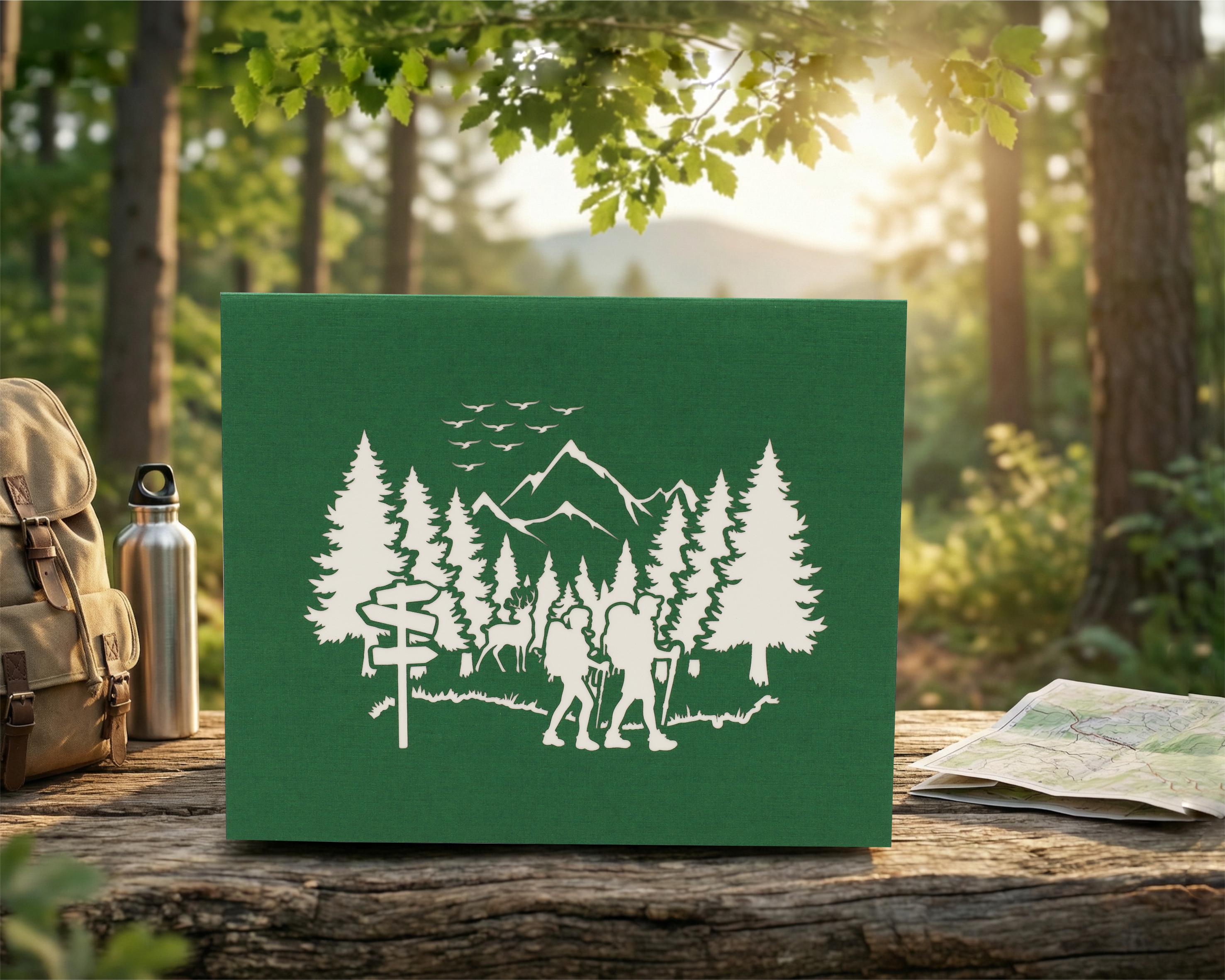 Grüne Klappkarte mit einem Wald, Bergen und 2 Wandersleuten in der Natur als Scherenschnitt. Die Pop Up Karte steht auf einer Bank in der Natur und im Hintergrund ist ein Wald.