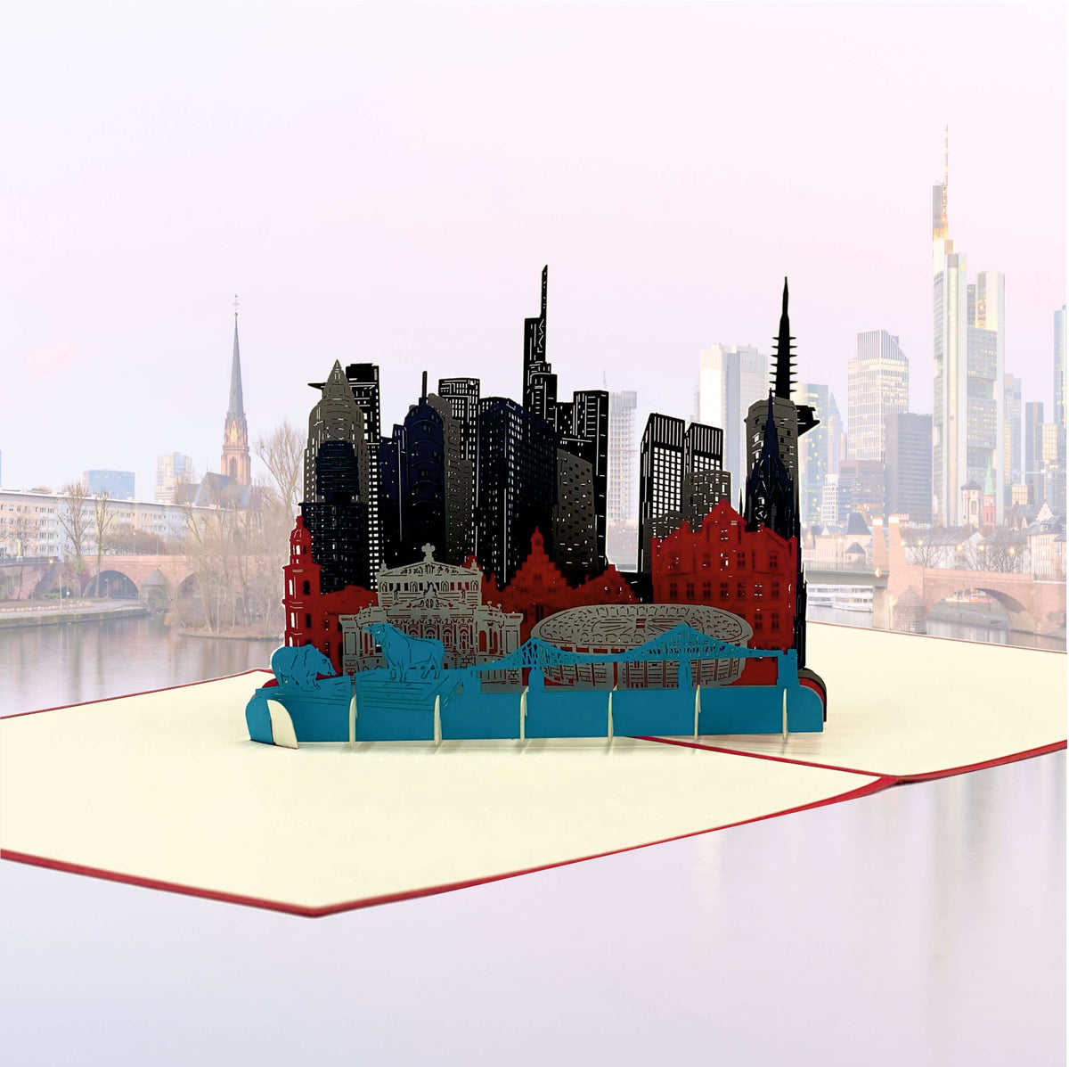 Aufgeklappte Pop-up-Karte mit der Skyline von Frankfurt vor einem echten Blick auf die Stadt am Main.