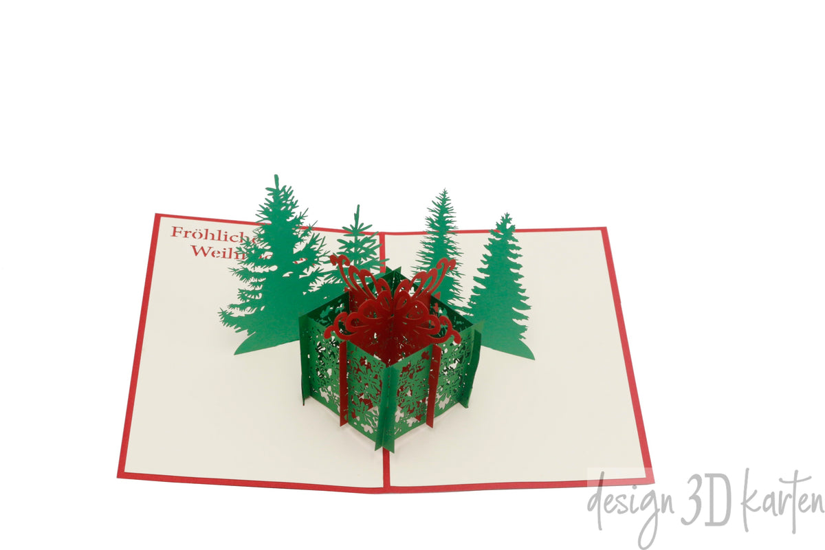Aufgeklappte Pop-up-Karte mit rotem Rand: eine grüne Geschenkbox mit roter Schleife zwischen Tannenbäumen, dazu der Schriftzug Fröhliche Weihnachten.
