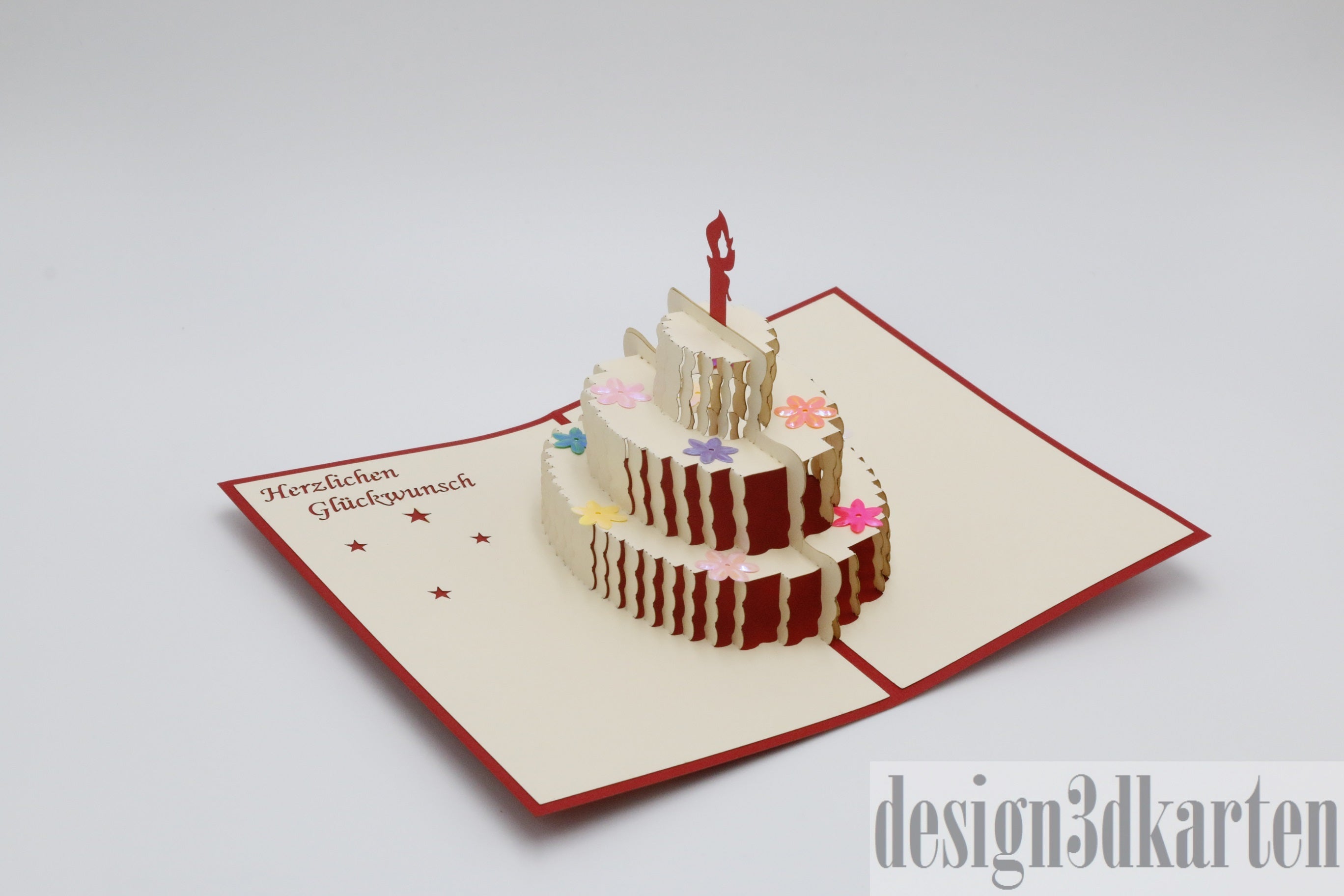 Aufgeklappte cremefarbene Pop-up-Karte mit roter Kante und einer dreistöckigen Geburtstagstorte mit Kerze, daneben der Schriftzug Herzlichen Glückwunsch.