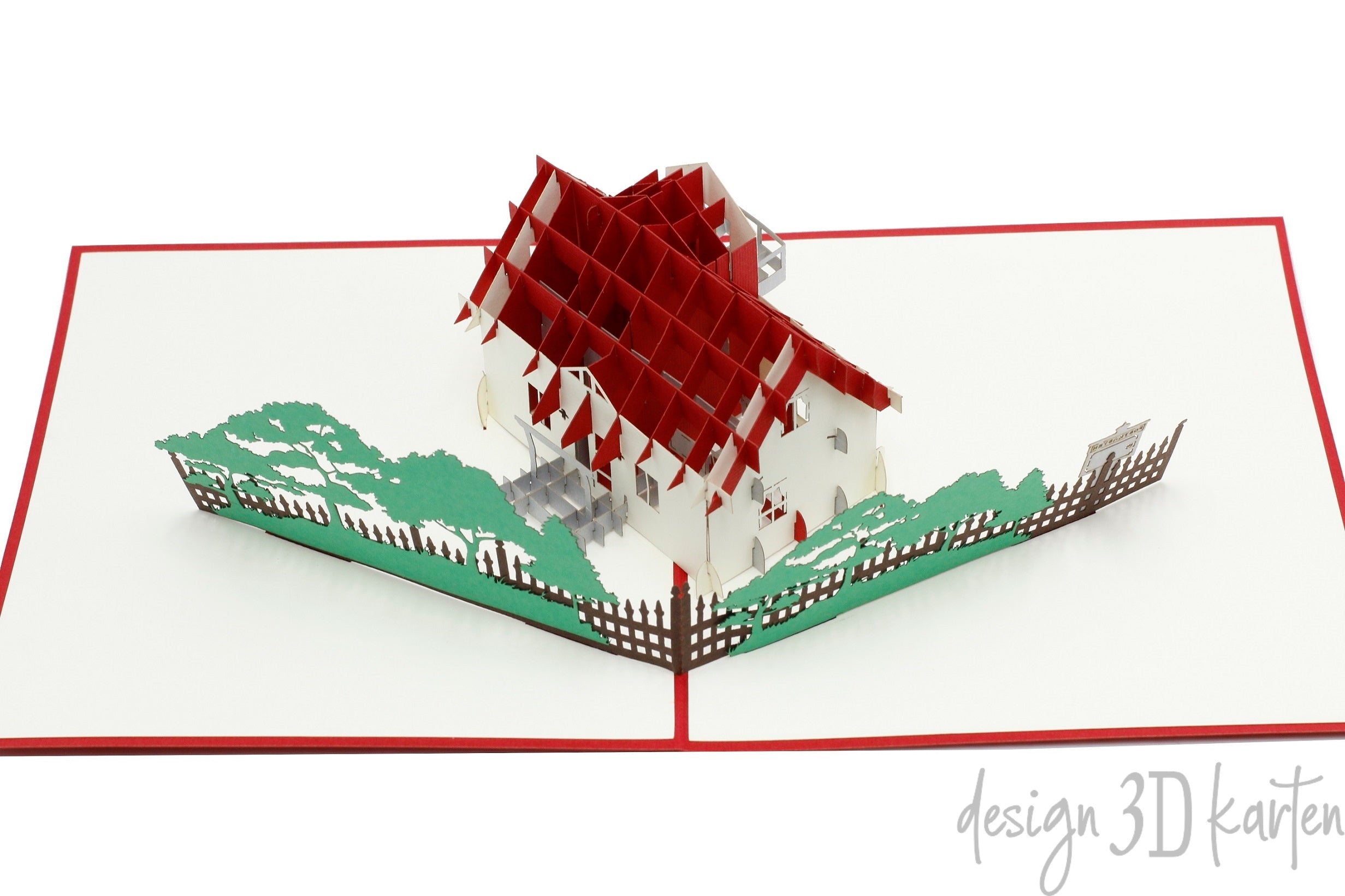 Aufgeklappte Pop-up-Karte mit Haus, Zaun und Bäumen, schräg von der Seite fotografiert.