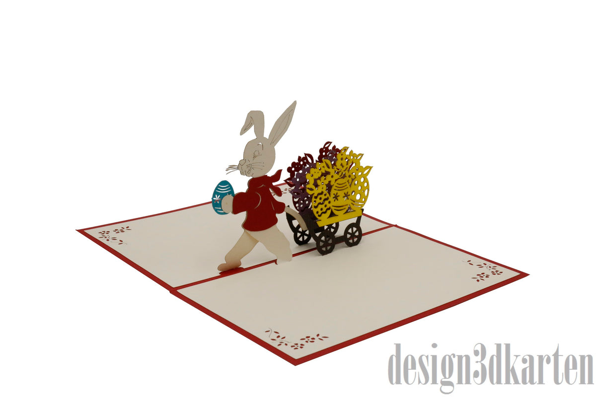 Aufgeklappte Pop-up-Karte mit einem Osterhasen im roten Mantel, der einen Handwagen voller gelber Blumen zieht.