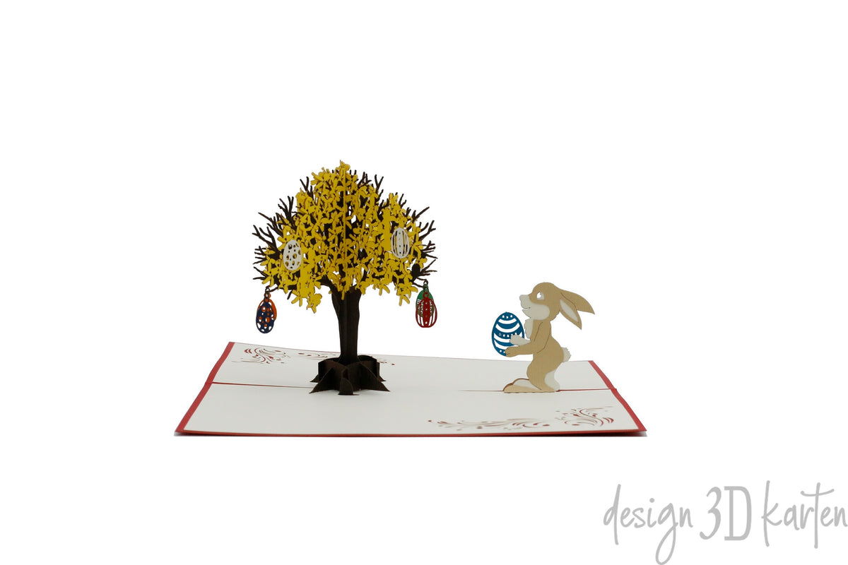 Aufgeklappte Pop-up-Karte mit rotem Rand: ein Osterhase mit blauem Ei neben einem gelb blühenden Goldregenbaum mit Ostereiern.