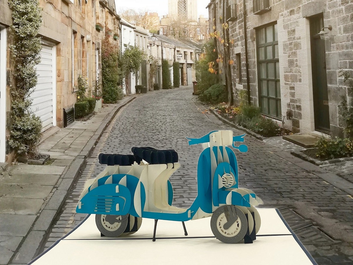 Pop-up-Karte mit blauer Vespa, aufgestellt in einer gepflasterten Altstadtgasse.