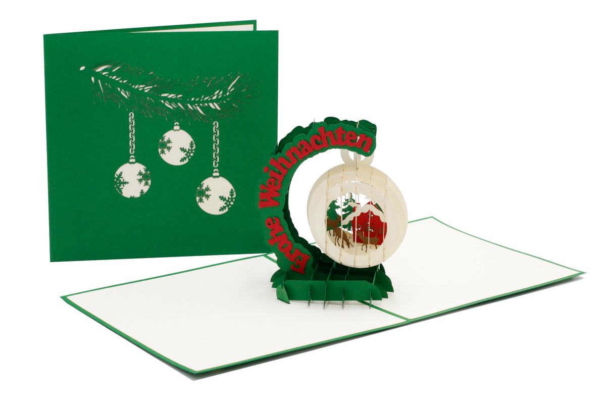 Aufgeklappte Pop-up-Karte mit einer Schneekugel mit Winterlandschaft und dem Schriftzug Frohe Weihnachten, daneben die grüne Kartenvorderseite mit Christbaumkugeln.